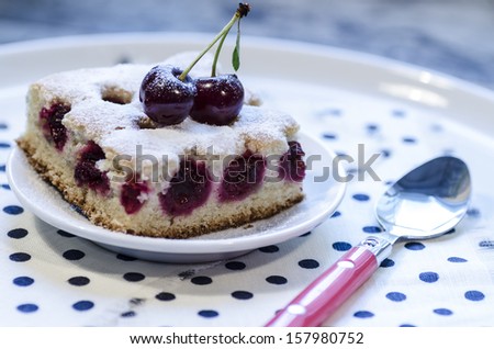 A slice of cherry pie on a linen napkin with polka dots. A cake decorated with fresh cherries. Next to a glass of juice. From the series 