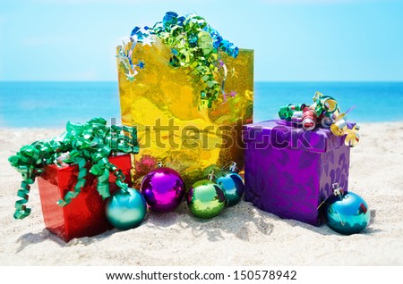 Gold gift bag and two gift boxes with Christmas balls on sandy beach in sunny day- holiday concept