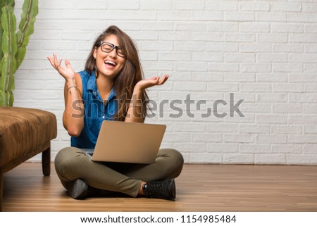 Portrait of young latin woman sitting on the floor laughing and having fun, being relaxed and cheerful, feels confident and successful. Holding a laptop.
