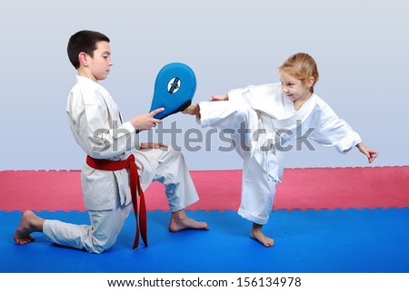 Little girl with a white belt beat leg on the simulator in the hands of a boy with a red belt