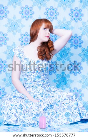 Vintage fashion styled redheaded woman sitting with an arm behind her head, wearing a blue patterned dress against a blue patterned backdrop (urban camouflage) - A bottle of pink soda in front of her