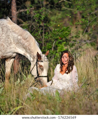 Attractive young woman in a vintage dress, sitting in long grass with her horse beside her.