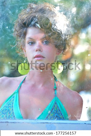 Attractive sultry woman with curly hair in a green dress looking through a dirty pane of glass that is in an old door.