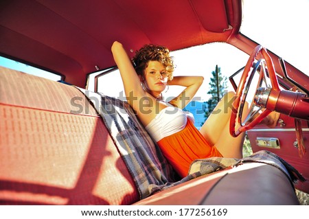 Attractive, sultry woman on a sunny day wearing an orange and white dress sitting in the drivers seat of an old red car with bench seats.