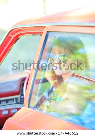 Attractive, smiling red headed woman in an old red car wearing sunglasses on a sunny day