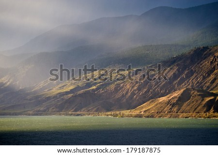 Sunlight hitting a green lake and the base of a mountain during a big storm