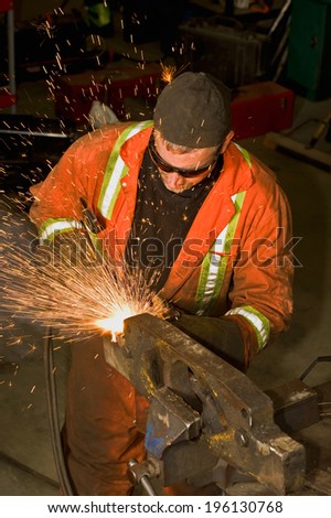 Heavy duty mechanic wearing orange overalls, grinding a piece of metal in a vise