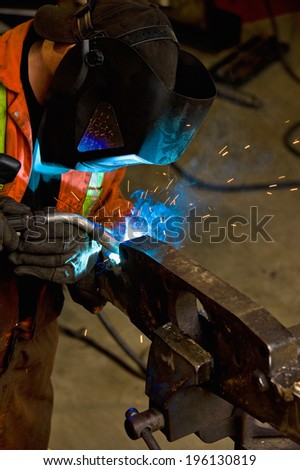 Mechanic wearing a safety mask and orange overalls, welding with an arc welder creating a blue flame and smoke