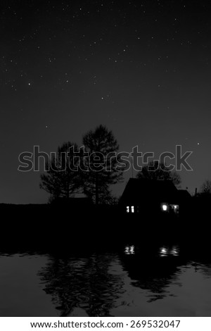 Night on the edge of the village. Dark night in the village. Black-and-white image. Reflection of the house and trees in a reservoir on the bank of the lake at night with stars in the clear sky