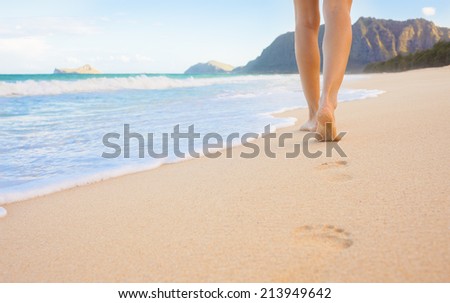 Beach travel - woman walking on sand beach leaving footprints in the sand. Closeup detail of female feet and golden sand on the beach, Hawaii, USA.
