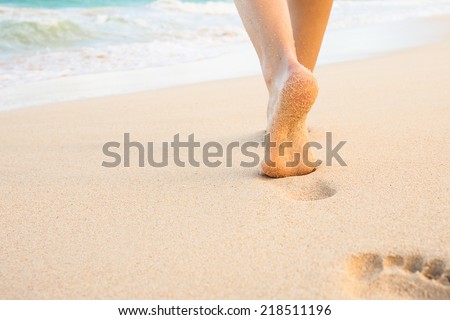 Beach travel - woman walking on sandy beach leaving footprints in the sand. Closeup detail of female feet and golden sand on beach in Hawaii
