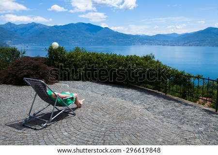 Woman in a green dress sits in a deck chair on a stone paved terrace with a view of Lake Maggiore, Italy