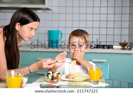 sister trying to feed her brother. he resists and repels fork with food. horizontal
