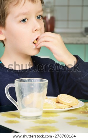 little boy eating cookies, sitting at the dinner table. biscuits on the table and a glass of milk. green and gray kitchen furniture in the background. vertical
