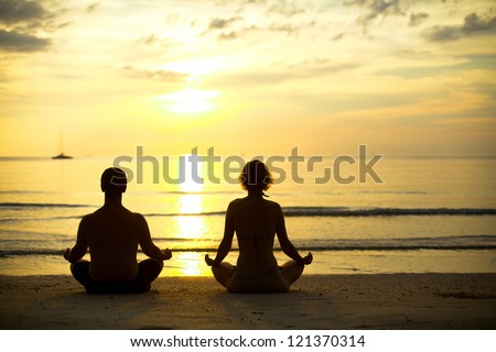 Yoga practice, a young couple sitting on the beach of the sea in the lotus position at sunset