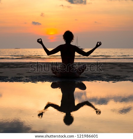 Yoga woman sitting in lotus pose on the beach during sunset, with reflection in water.
