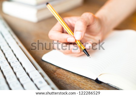 Hands writes a pen in a notebook, computer keyboard and a stack of books in background