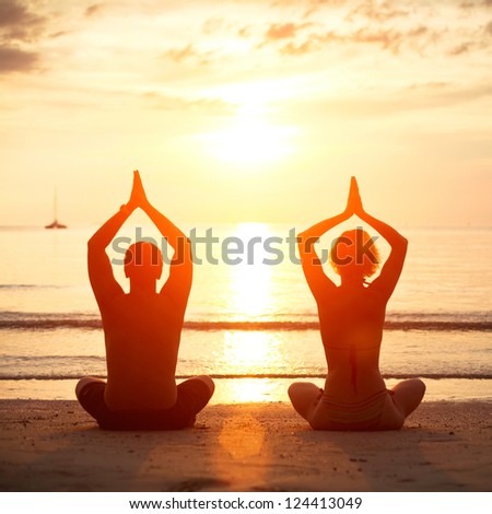 Yoga practice, young couple sitting on the beach of the sea in the lotus position at sunset.
