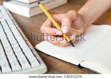 Young woman hand writes a pen in a notebook, computer keyboard and a stack of books in background