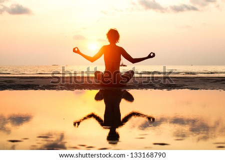 Yoga woman sitting in lotus pose on the beach during sunset in bright colors. (with reflection in water)