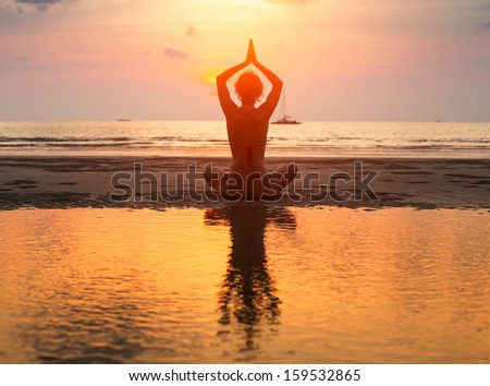 Yoga woman sitting in lotus pose on the beach during sunset with reflection in water.