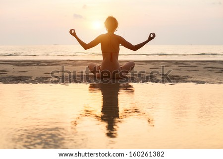 Yoga woman sitting in lotus pose on the beach during sunset (with reflection in water)