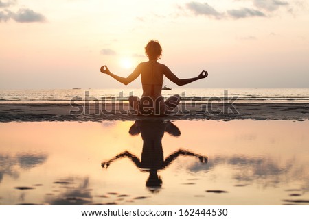 Yoga woman sitting in lotus pose on the beach during sunset, in bright colors, with reflection in water.