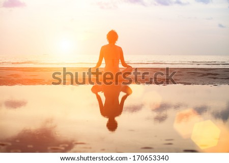 Yoga woman sitting in lotus pose on the beach during sunset, with reflection in water.
