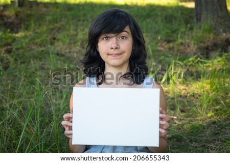 Lovely teen-girl holding clean white sheet paper, in the park (banner for your message)
