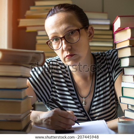 Intellectual woman writes on a notebook in a room with lots of books (loking at the camera)