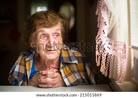 Old lonely woman sitting at a table near the window in his house. Old age. Loneliness.
