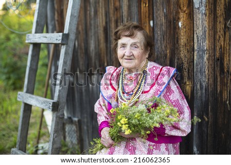 Old woman in ethnic clothes with wild flowers in their hands in the yard of his rural home.