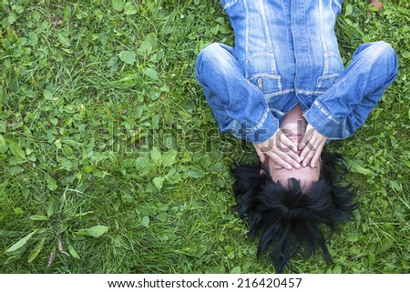 Young woman in a denim jacket lying on the grass and closes his eyes with his hands (with space for text)