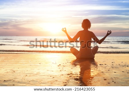 Yoga woman in the lotus position sitting on a sea beach during sunset.
