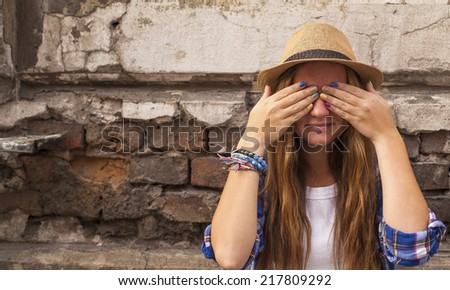 Hipster girl on a street in the city closes his eyes with his hands on the brick wall background.