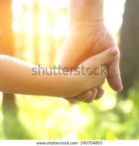Adult holding a child\'s hand, close-up hands, nature in background.