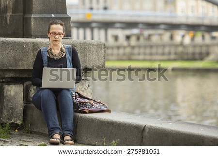 Young woman typing on laptop sitting on embankment of river in the old town.