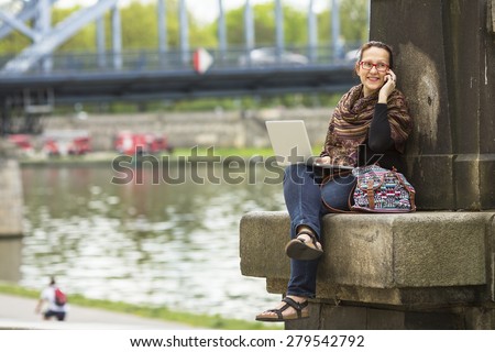 Young woman with laptop sitting on the embankment of the river in the old town talking on a cell phone.
