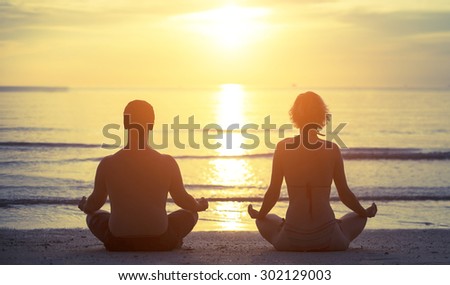 Silhouettes of a young couple sitting in the yoga Lotus position on the banks of the sea during sunset.