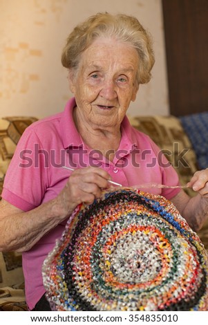 An elderly woman knits crochet rug.