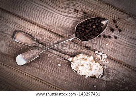 Vintage effect image of tarnished silver spoons filled with salt crystals and black peppercorns, over rough wood background.