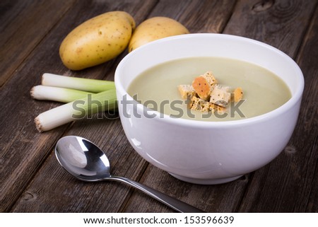 A Bowl Of Leek And Potato Soup With Bread Croutons, Over Old Wood Table With Fresh Leeks And Potatoes Alongside