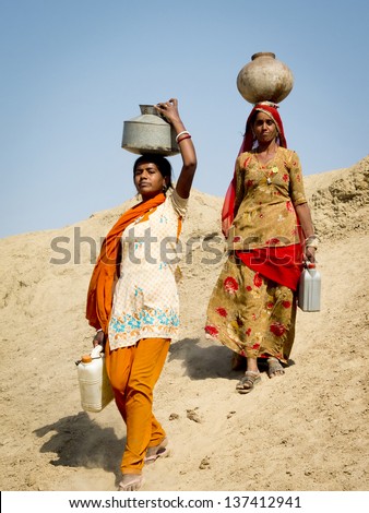 Jaisalmer, India - March 8: Close-Up Portrait Of Two Women Carrying Water In The Desert , On March 8, 2013, Jaisalmer, India. They Are Walking On A Dune With Heavy Jugs On Their Heads.