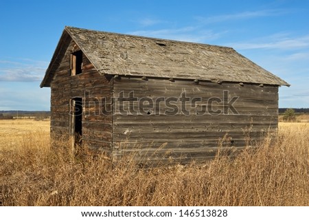 An old wooden granary in fall with dry weeds and grass in the foreground and a harvested field in the background
