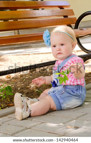 beautiful little girl in denim overalls and a plaid shirt sitting on the ground near a bench in the park