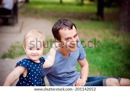 little girl in a polka dot dress plucks his dad behind the ear in the park