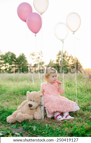 baby in a pink dress sitting on a bench in the woods with teddy bear and balloons and eating candy