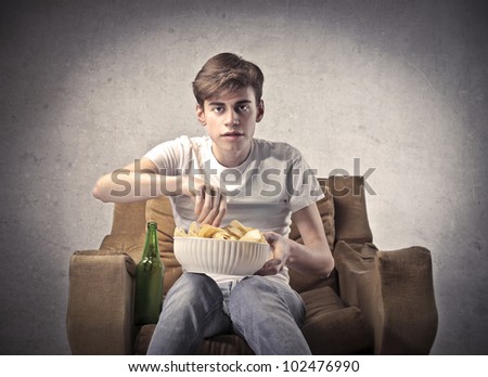 Young man sitting on an armchair with a beer beside him and a bowl of chips on his knees