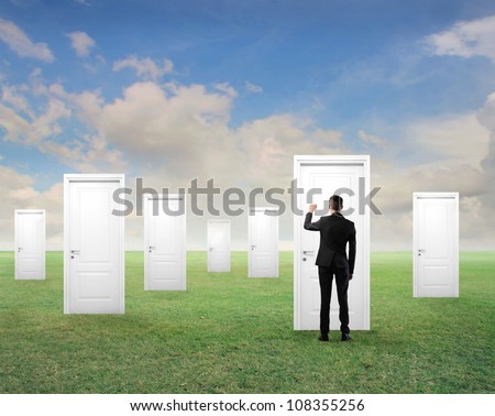 Young businessman knocking at one of many white doors on a green meadow