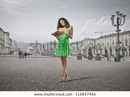 A beautiful brown girl is reading a book while is standing in the center of a town